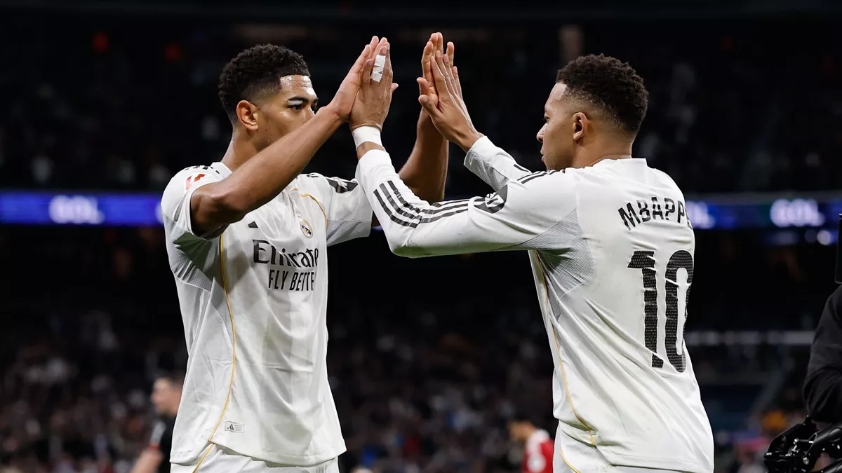 Los jugadores del Real Madrid Jude Bellingham, a la izquierda, y Kylian Mbappé, de espaldas con el dorsal 10 a la derecha, chocan las dos manos en el aire celebrando en el campo de juego durante un partido con público en las gradas de fondo.