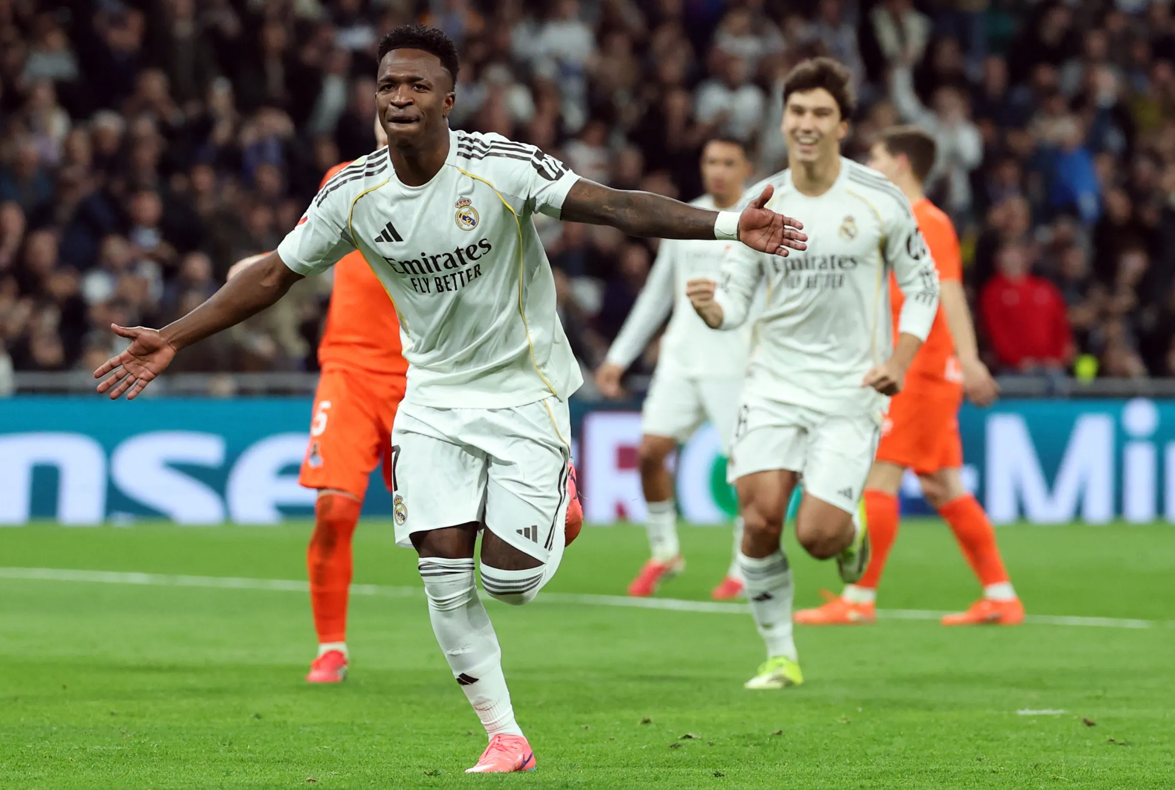 El delantero del Real Madrid, Vinicius Junior, corre por el campo con los brazos abiertos celebrando un gol, mientras su compañero de equipo sonríe y corre detrás de él para abrazarlo, en un estadio lleno de aficionados borrosos al fondo.