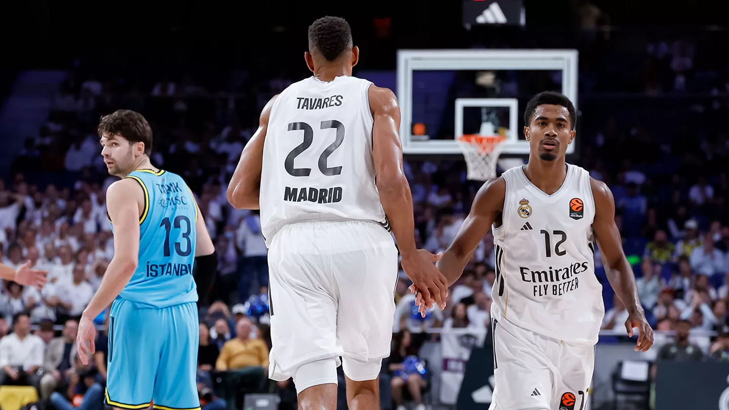 El pívot del Real Madrid Edy Tavares, de espaldas con el dorsal 22, choca la mano con su compañero Theo Maledon (dorsal 12) en la cancha de baloncesto, mientras un jugador del Fenerbahce con camiseta azul observa a la izquierda bajo la canasta.